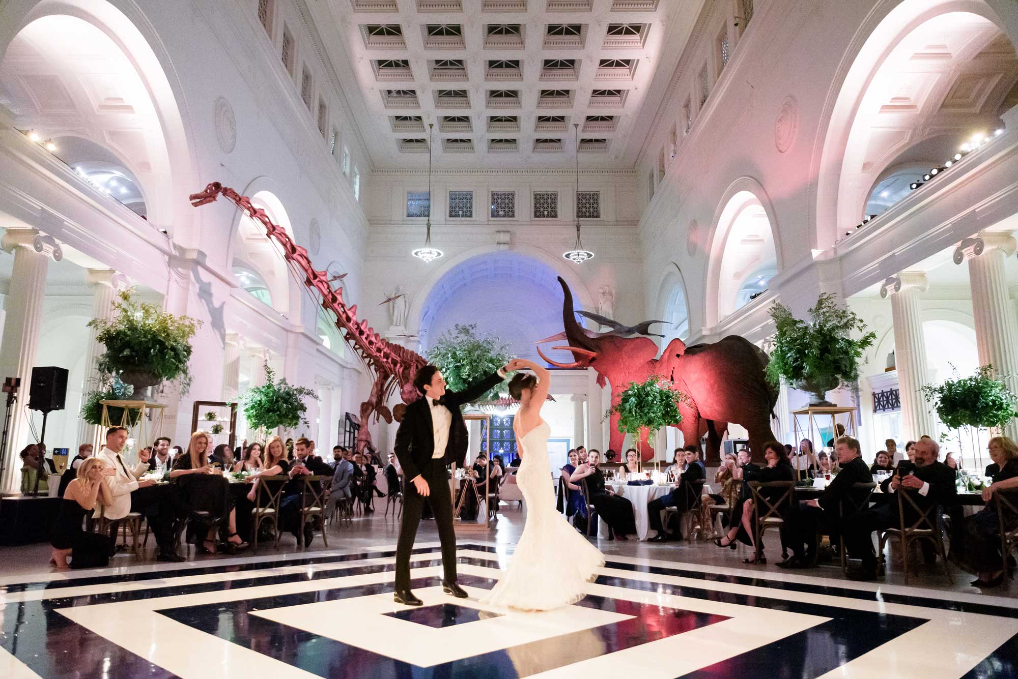 A couple shares their first dance in the center of Stanley Field Hall on a black and white dance floor. Wedding guests watch from their seats and elephants and Máximo the Titanosaur are visible in the background.