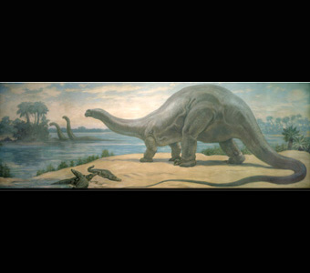 An apotasaurus standing on a sandy ground, while three much smaller crocodile-like creatures stand below. Two other long-necked dinosaurs are in the water in the background, reaching their heads toward tall trees.