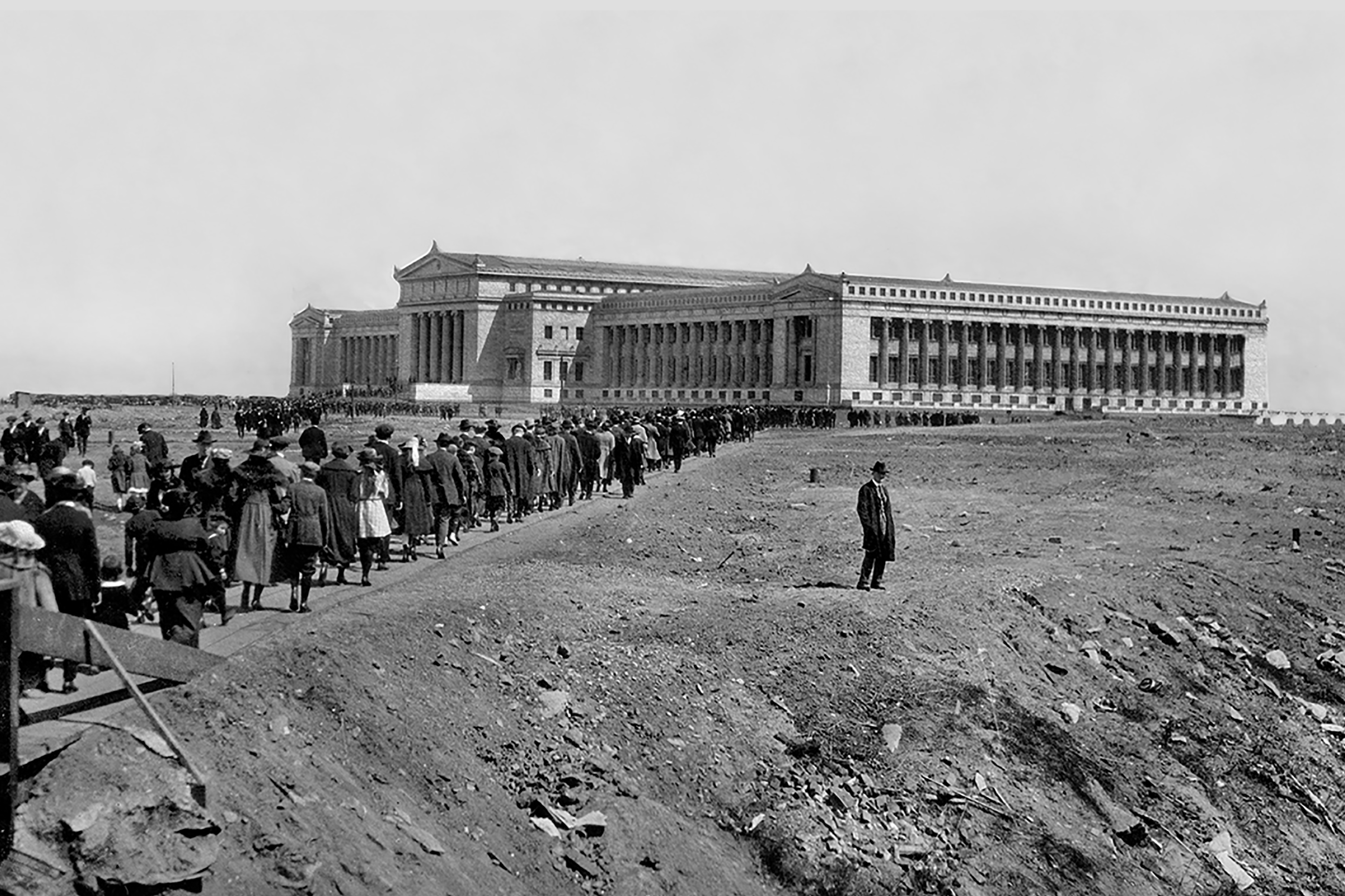 Crowds cross a wooden bridge and approach the Field Columbian Museum on opening day. A lone man stands to the side. The entire exterior view of the Museum’s north and west facades are visible in the background.