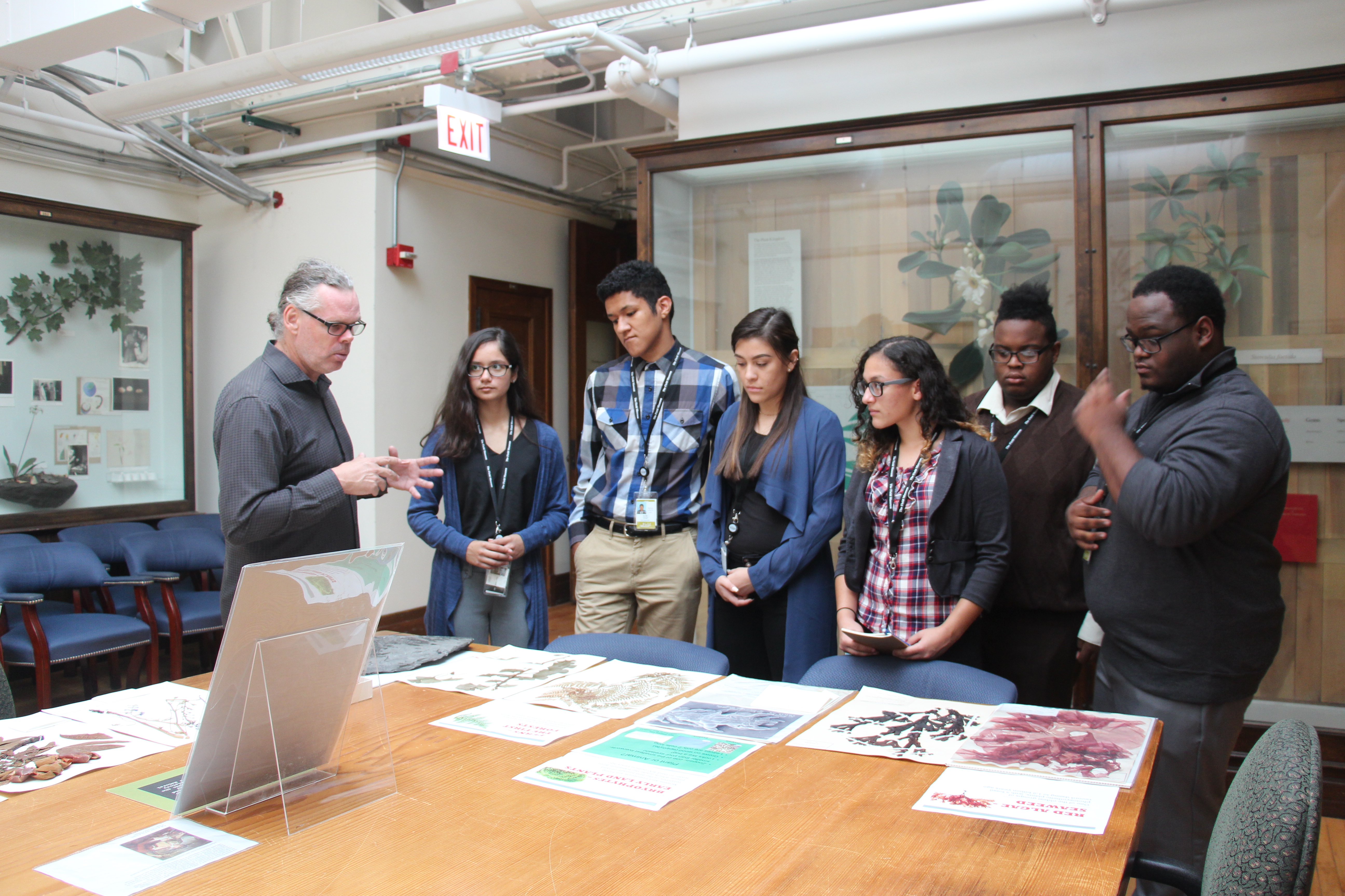 A scientist shares information about the botany collection at the Field Museum with a group of students in a classroom. Images of various plant specimens are laid out on a table as the scientist holds his hands out and lectures. Several cases of wax plant models visible in the background.