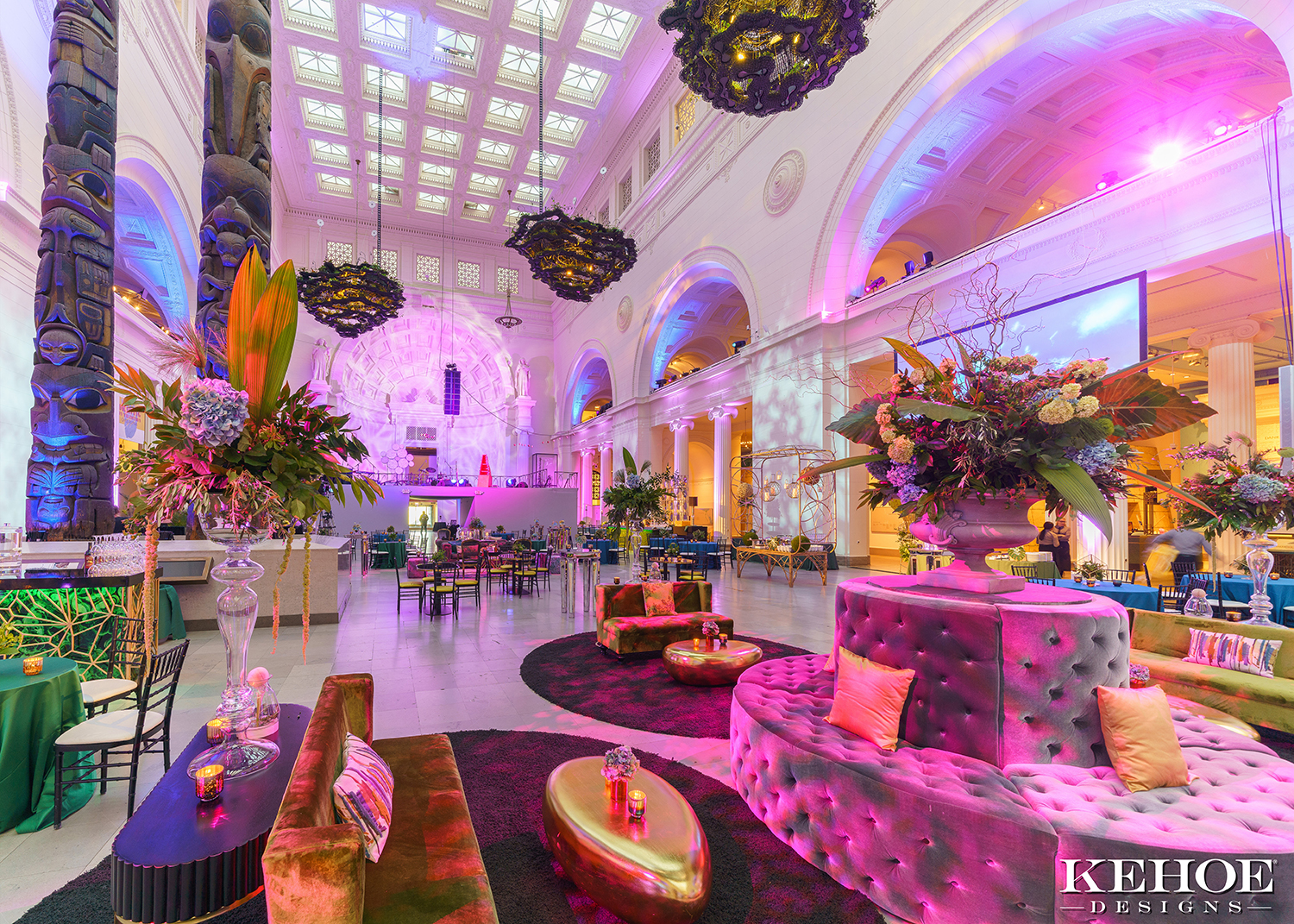 Warm, pink light fills Stanley Field Hall at the Field Museum, which is set up for an event with colorful tufted couches, rugs, and gold coffee tables. Floral arrangements decorate the space.