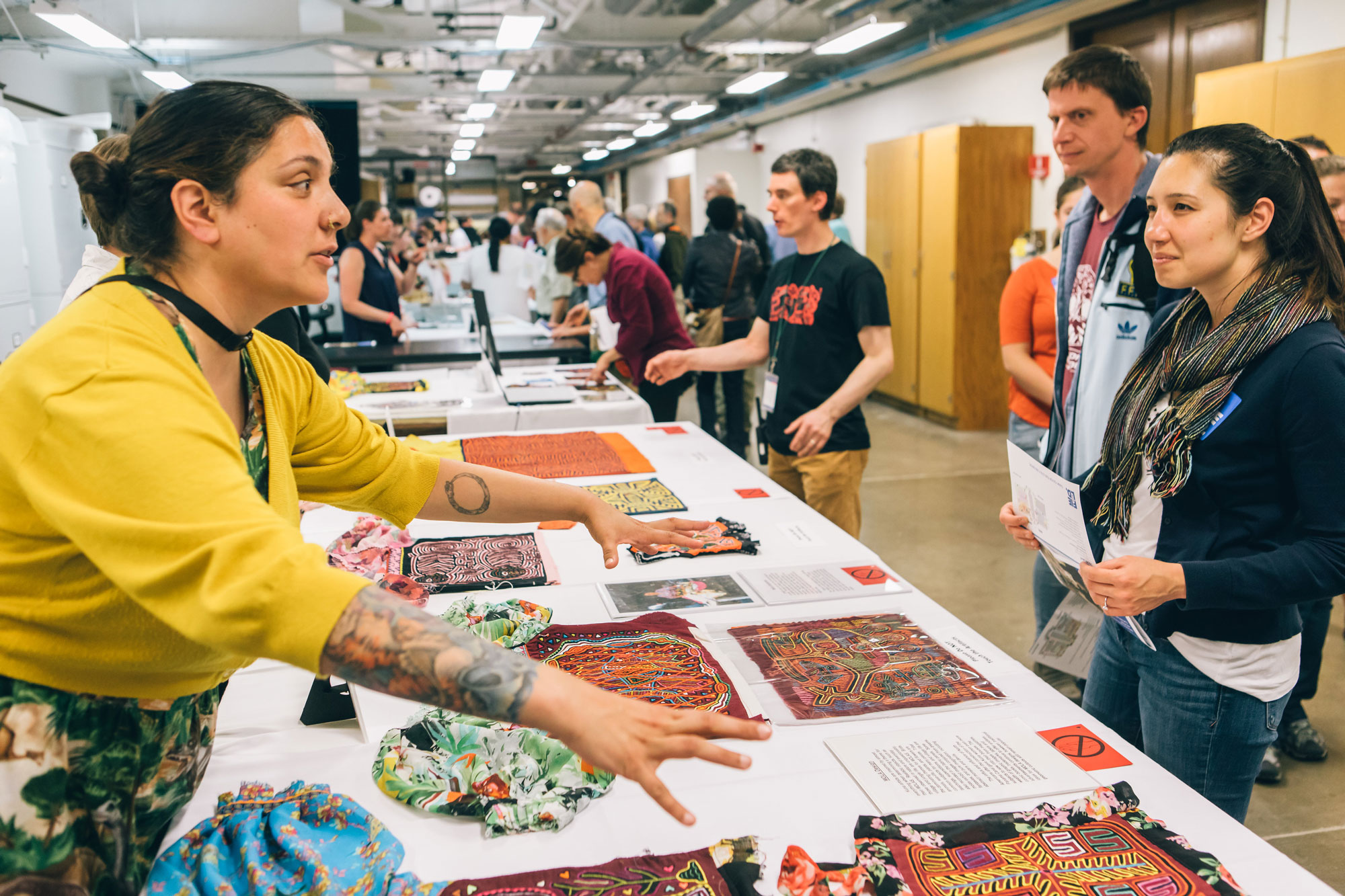 A woman stands behind a table featuring Native American textiles. She leans forward with her arms and hands outstretched as she talks to guests standing in front of the table.