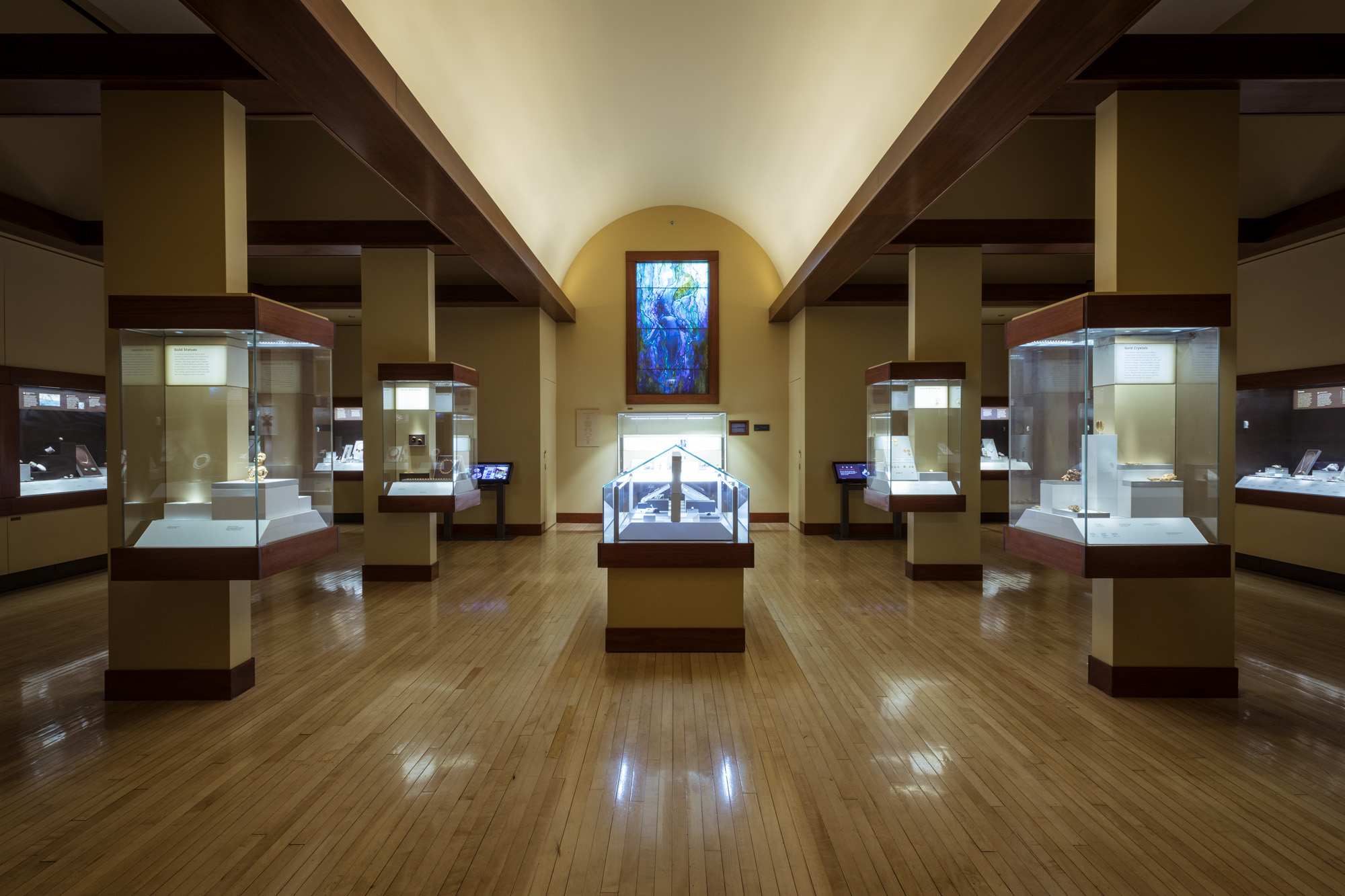 View of the Grainger Hall of Gems from the entrance, with many display cases visible. A stained glass window featuring a mermaid is visible in the center.