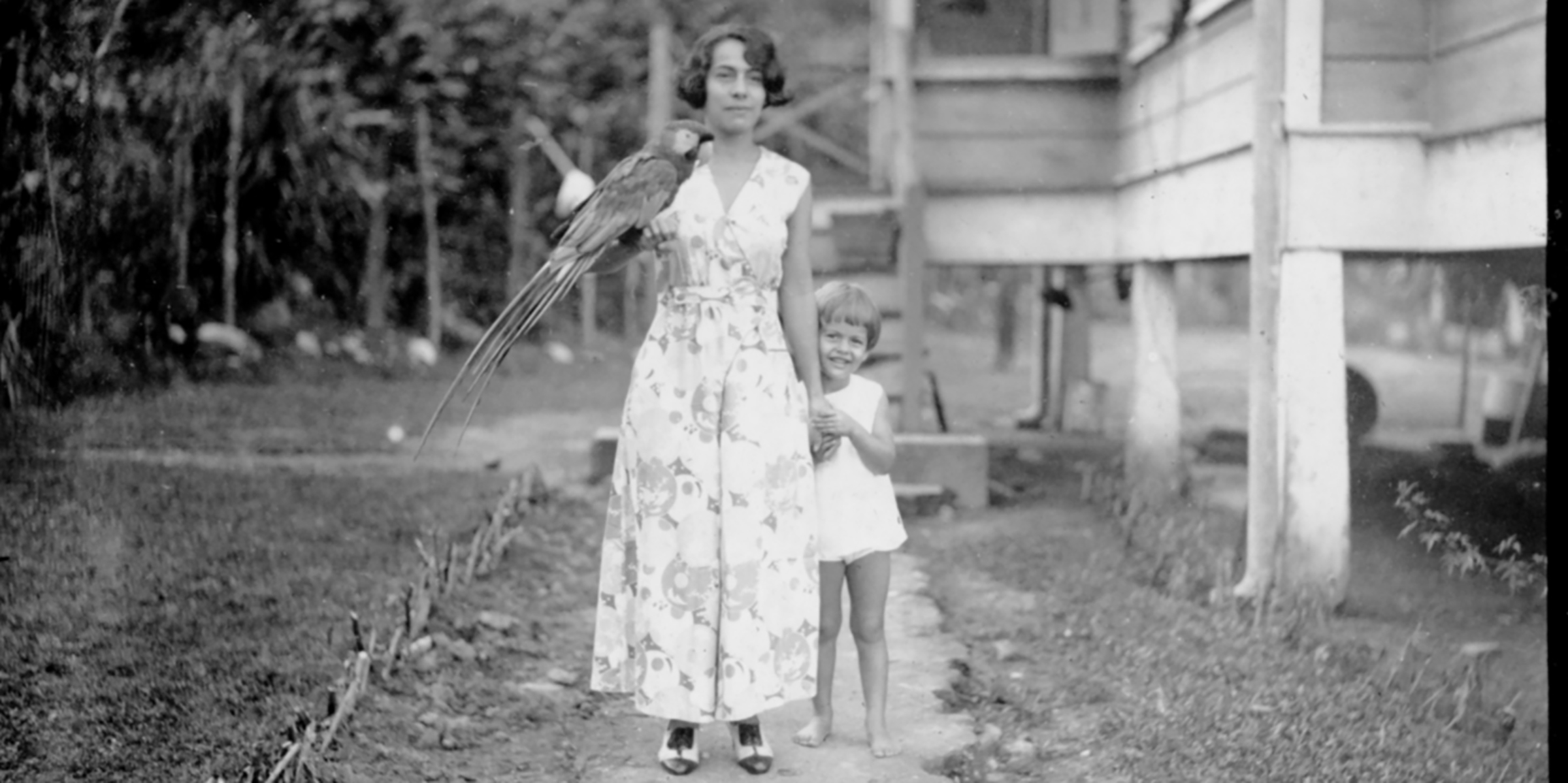 Black and white photograph of a woman in a floral dress with a large bird on her hand, and a small child smiling and standing at her side
