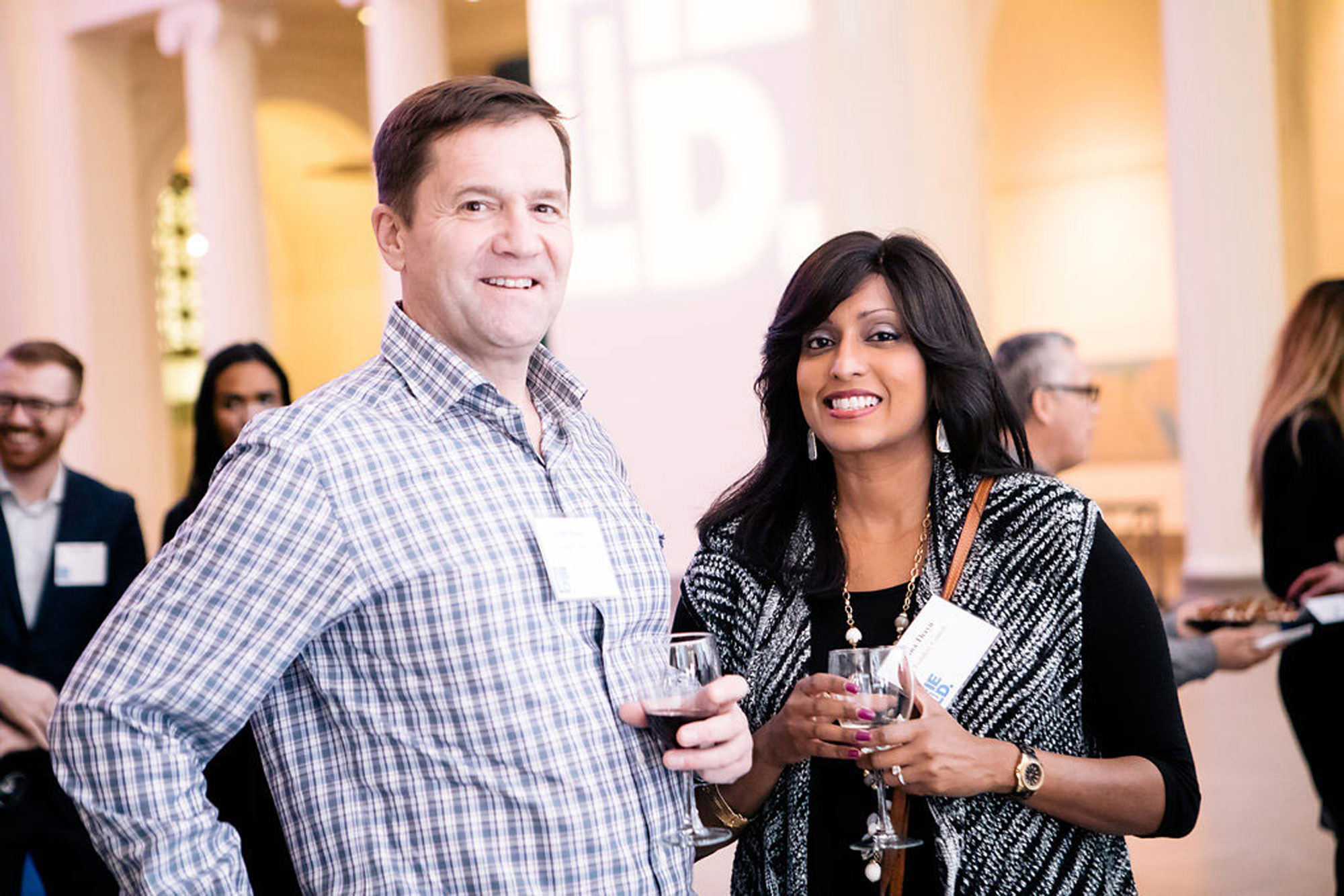 Two President’s Leadership Council members meet in Stanley Field Hall. They are smiling at the camera while holding wine glasses. Other council members stand in the background.