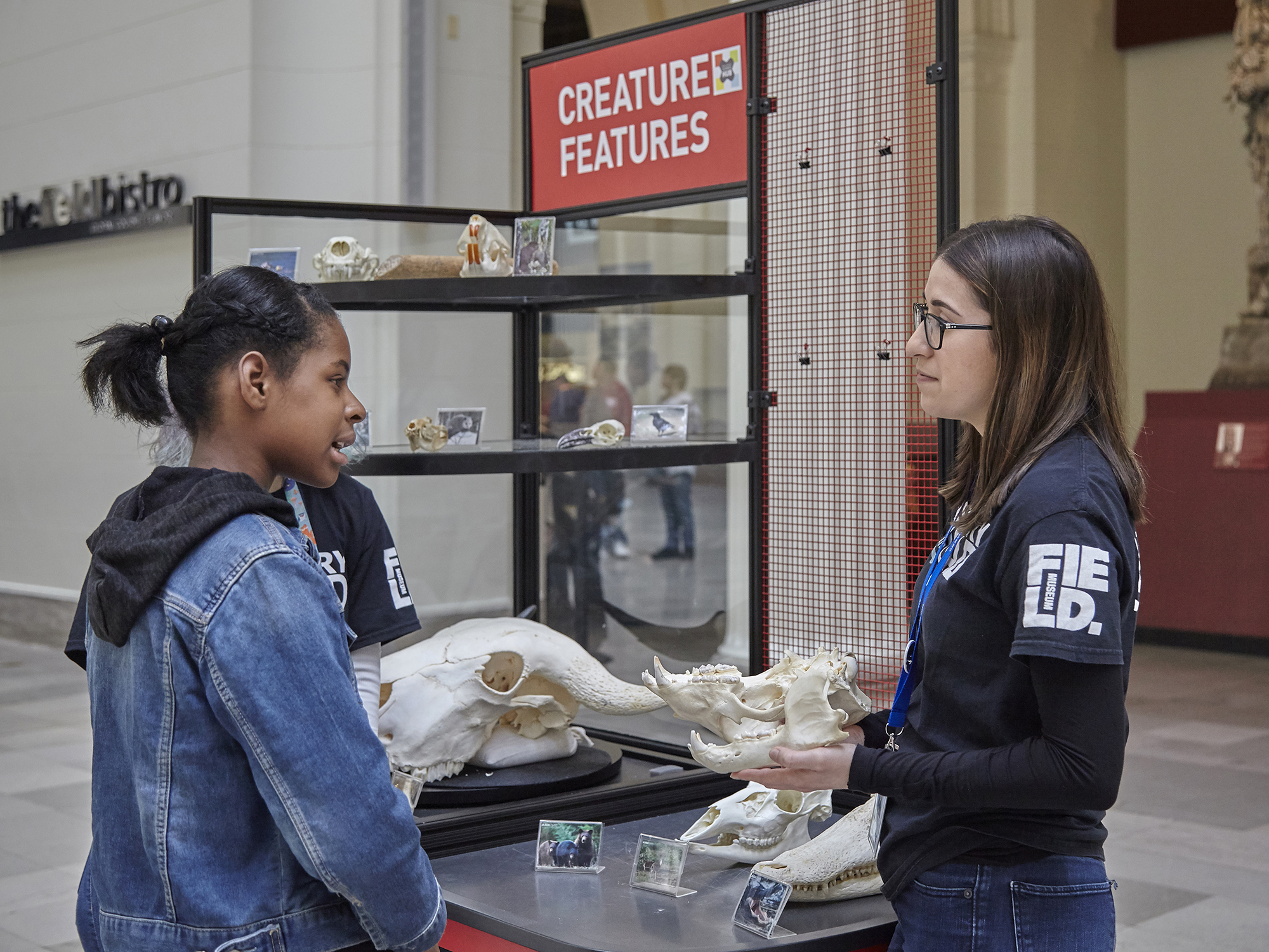 A young woman asks questions about an animal skull in front of a cart labeled Creature Features.