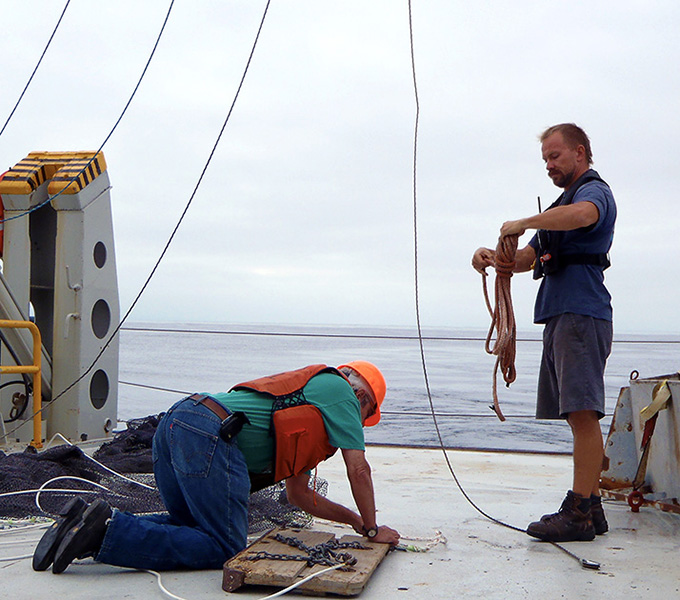 Chris Jones and the marine technician attaching the otter trawl for our first collection on the cruise.  The wood otter boards in the image are paired, and they are positioned such that the hydrodynamic forces acting on them as the boat moves forward, pushes them outwards; these forces prevent the mouth of the net from closing. They also act like a plough, mucking up the seafloor. This creates a turbid cloud, which scares fishes and other creatures toward the mouth of the net.