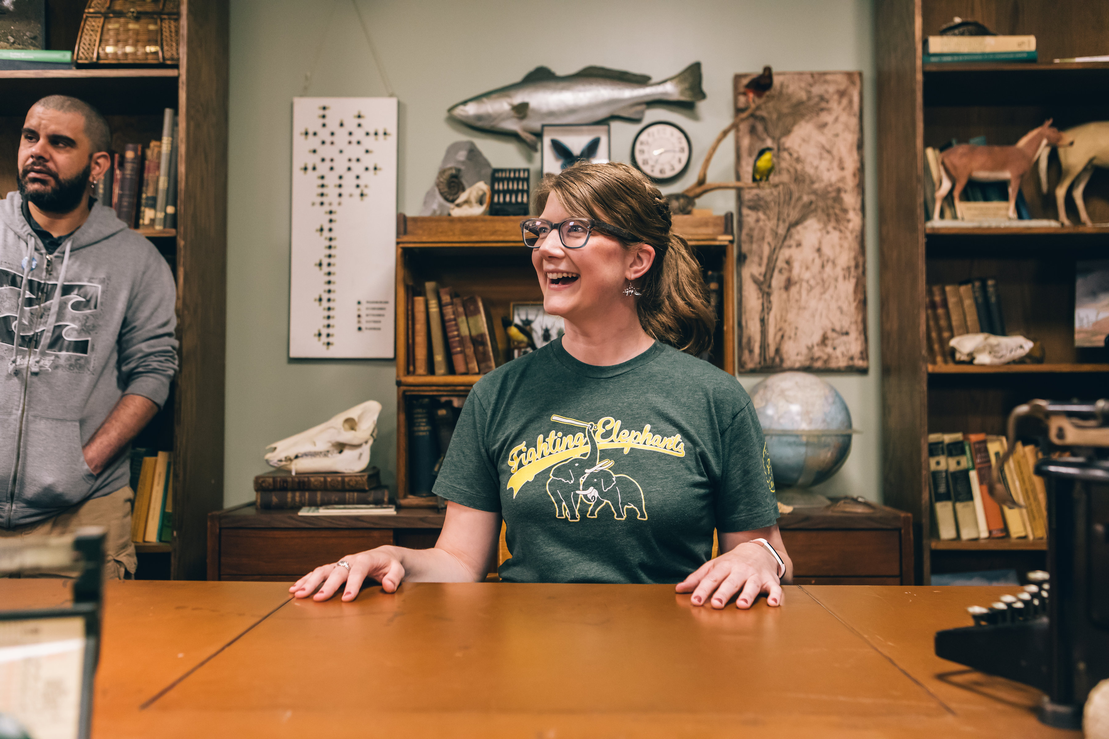 Emily Graslie smiles and looks to her right, while sitting at a desk. Books and specimens are visible in the background.