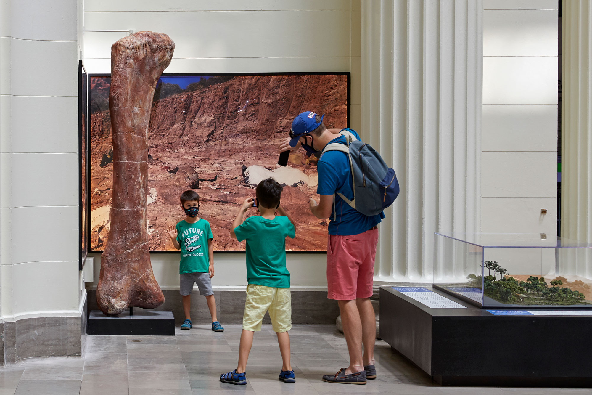A young boy wearing a face mask poses for a photo next to a dinosaur bone that's twice his height, in front of a mural of a rocky landscape. Another young boy and a man take his photo.