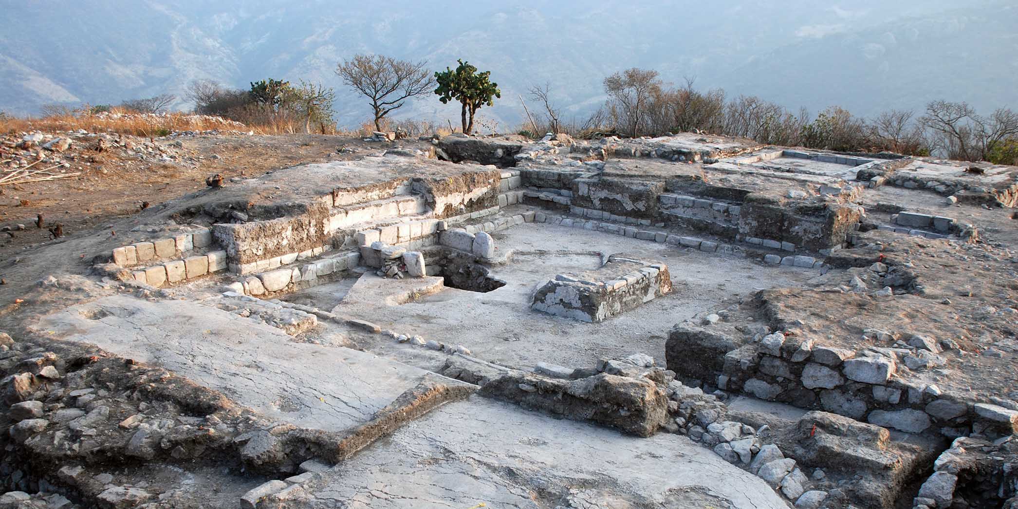 Archaeological ruins in the outline of a house, with trees and mountains in the background