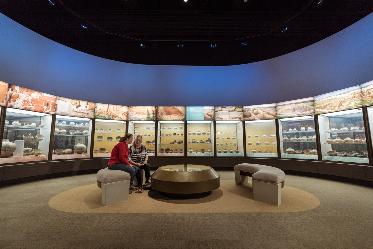 Two women sit on a bench in a circular space, surrounded by glass cases filled with pottery in a gallery in Robert R. McCormick Halls of the Ancient Americas.