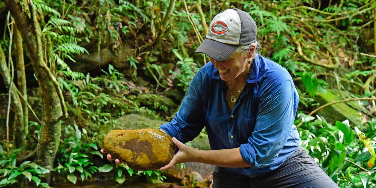 Woman wearing a baseball cap and blue shirt in a rainforest-like setting, holding a large rock