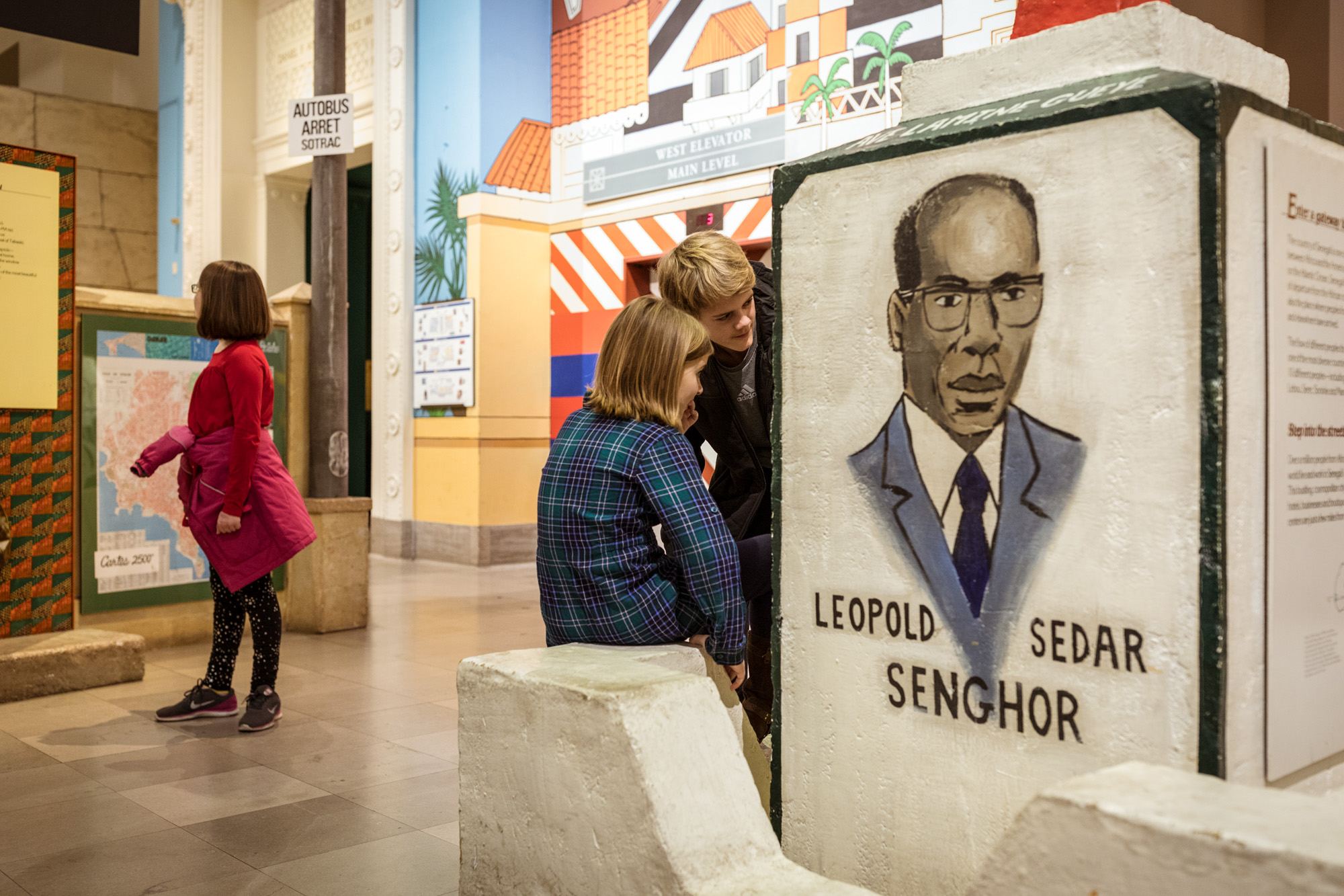 Three children explore near the entrance to the Africa exhibition. In the foreground is a sign with a drawing of the bust of a man wearing glasses and a suit and tie with the words “Leopold Sedar Senghor” written underneath.