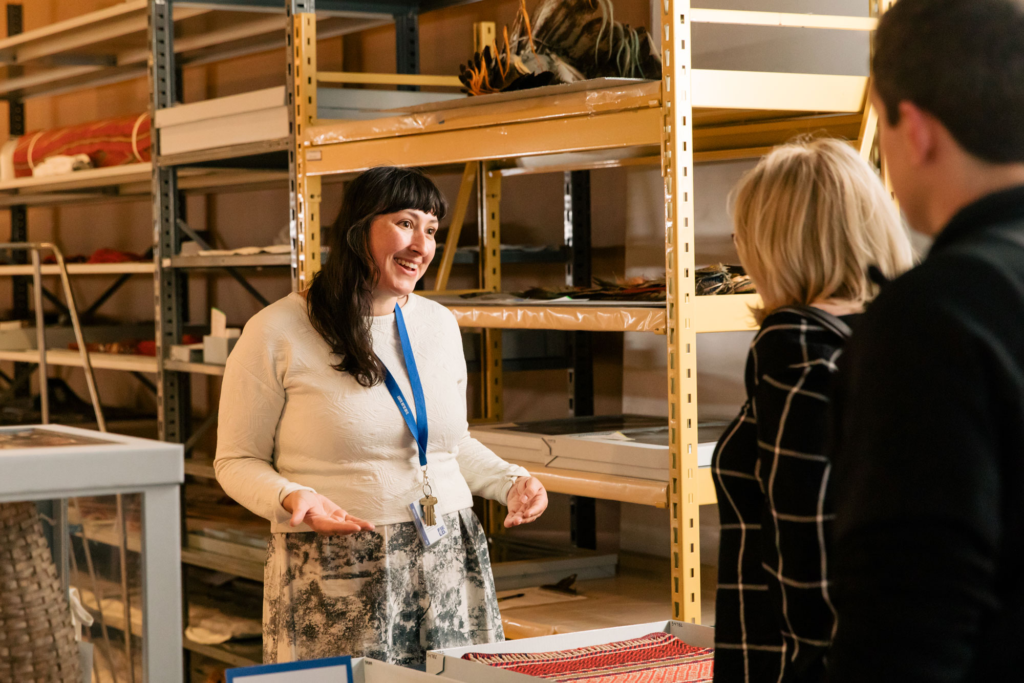 A woman behind a table speaks to two people. There are cultural items on the table and on many shelves behind the woman.