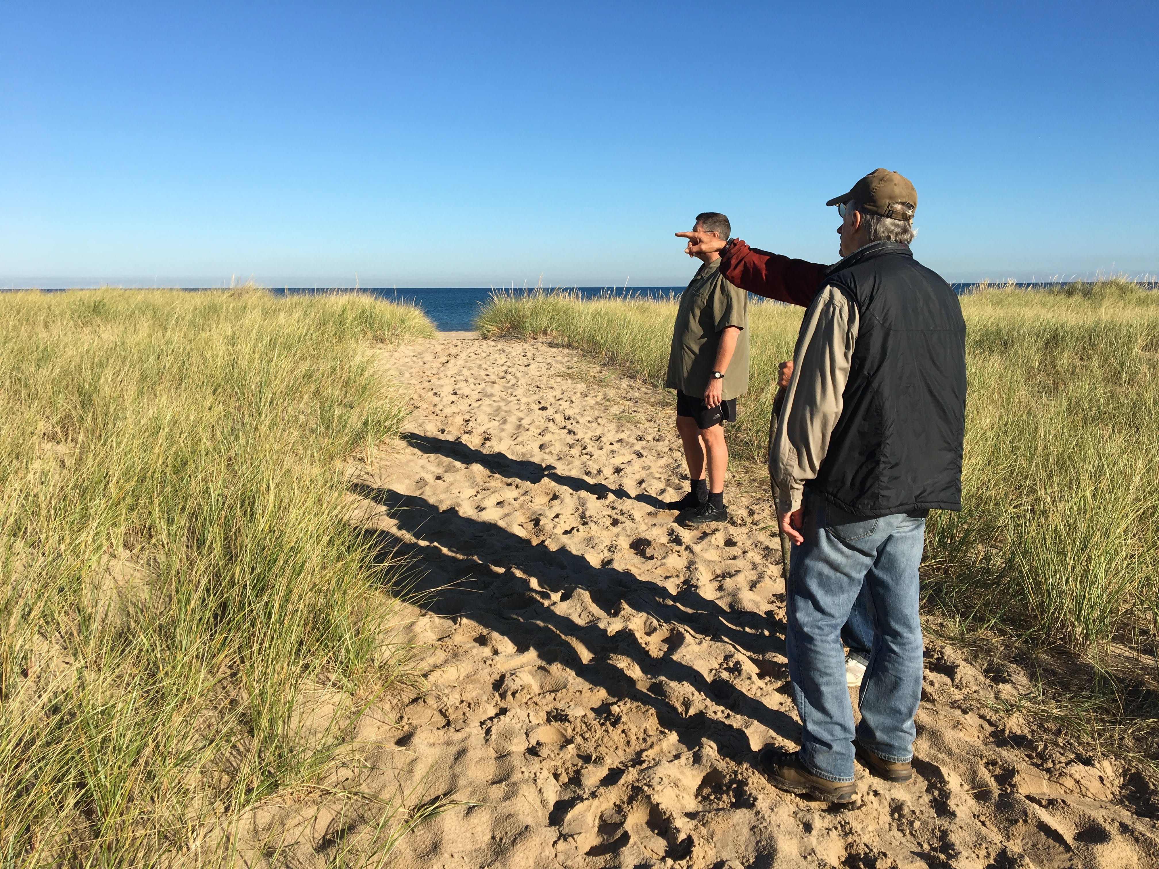 Three men hike a sandy trail in Dune Acres, IN. They all look to the left as one holds his arm out to point to something in the distance.