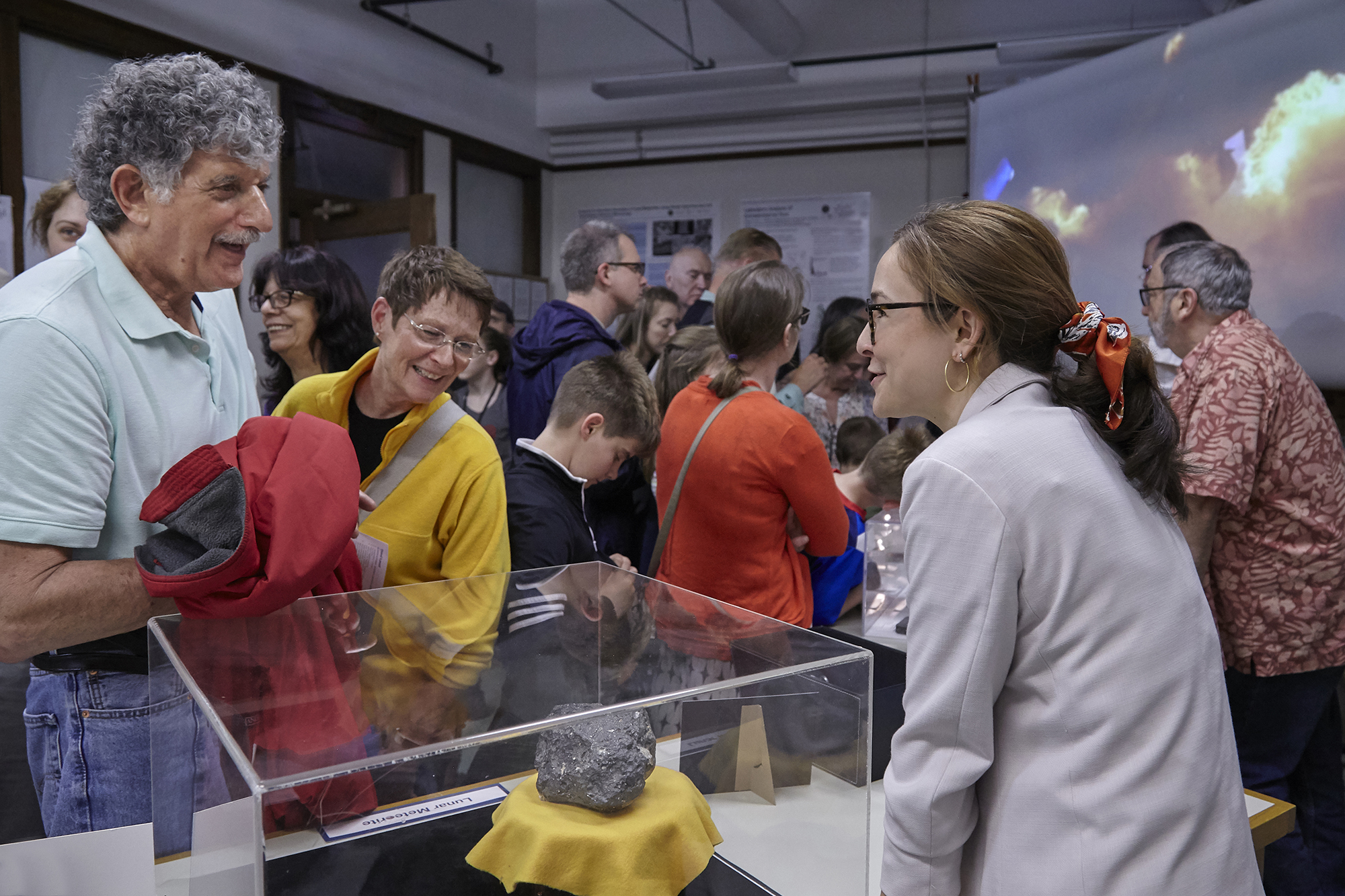 A scientist presents a meteorite to a crowd of people. She stands behind a desk with the meteorite encased in glass. People in the background crowd around another, unseen table.
