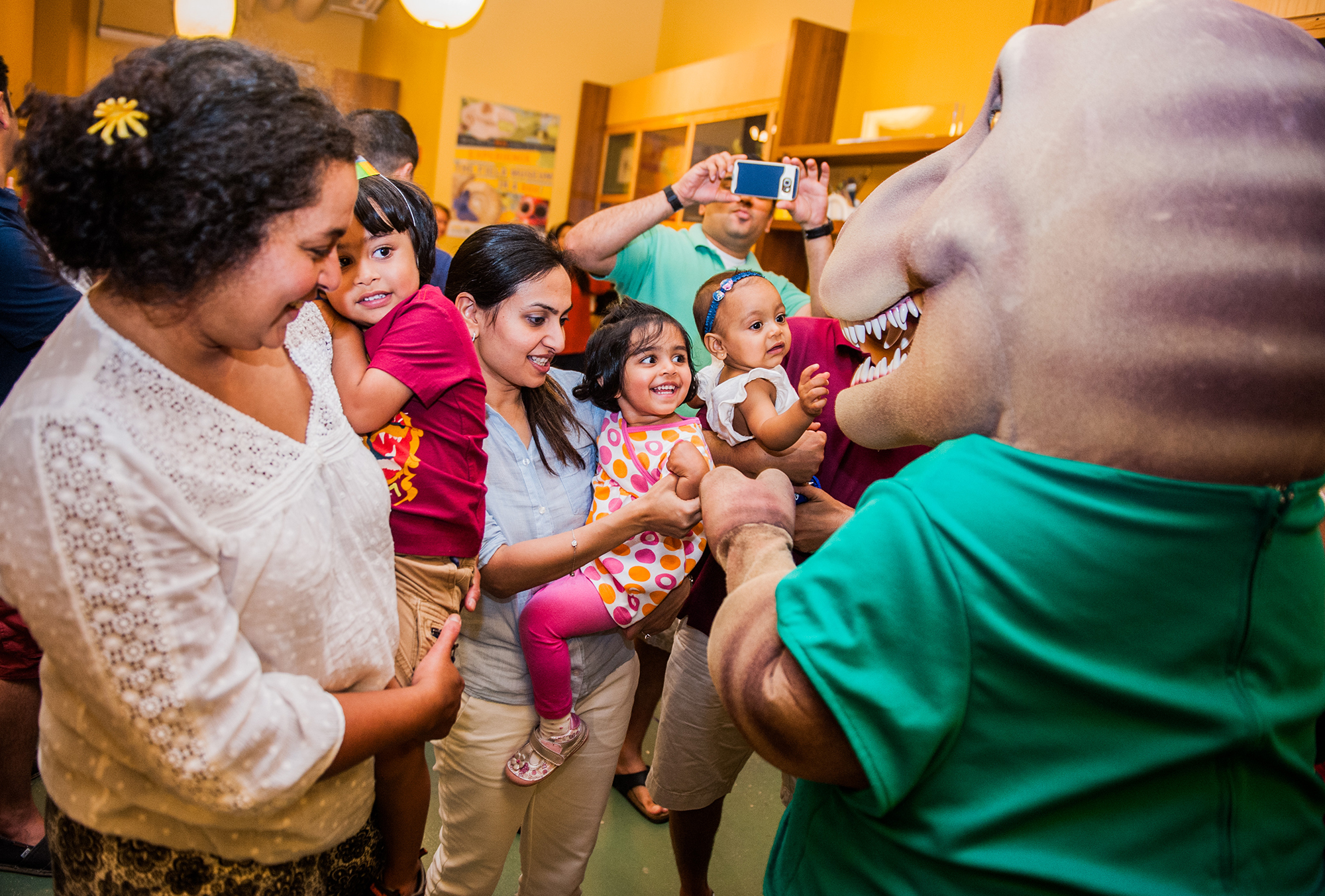 A group of three toddlers and their guardians interact with a T. rex mascot. The toddlers, held by their guardians, smile at the mascot. A man stands in the background taking a photo of the scene; the camera blocks his face.