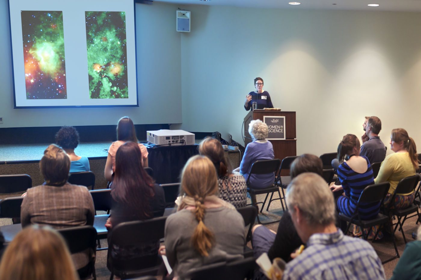 A crowd gathers to hear a speaker in Lecture Hall II. People sit in chairs facing a stage. A speaker stands at a podium, and a projector casts an image of outer space onto a screen against the wall.