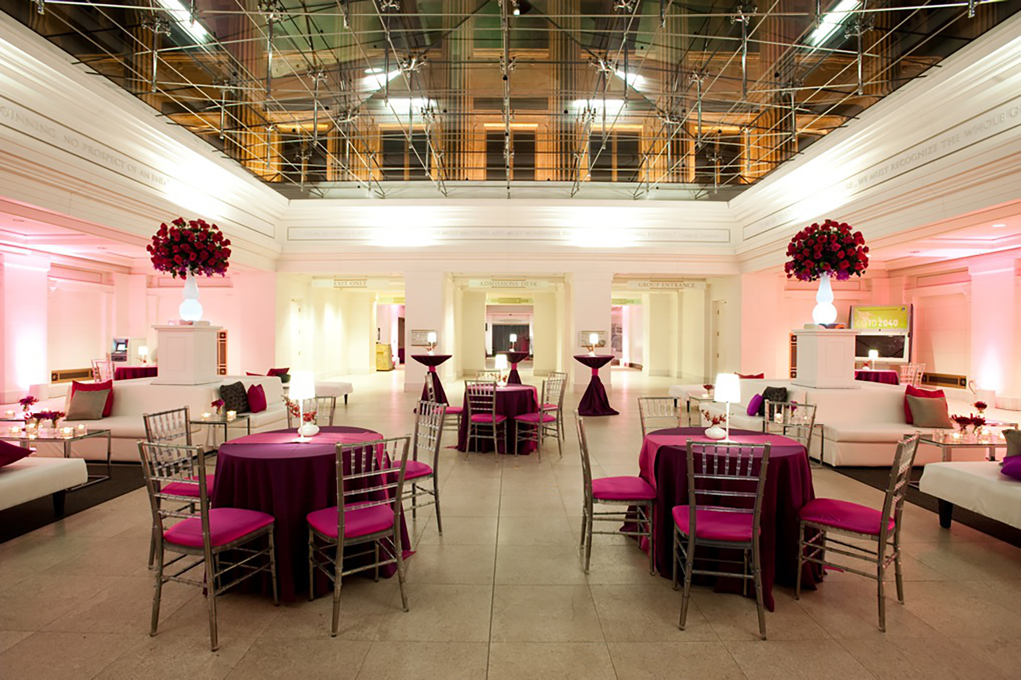 East Atrium space set up for an event with tall cocktail tables, small tables with five chairs eat, and white couches. Floral arrangements sit on while pillars behind the couches. A glass ceiling reveals the east facade of the Field Museum at night.