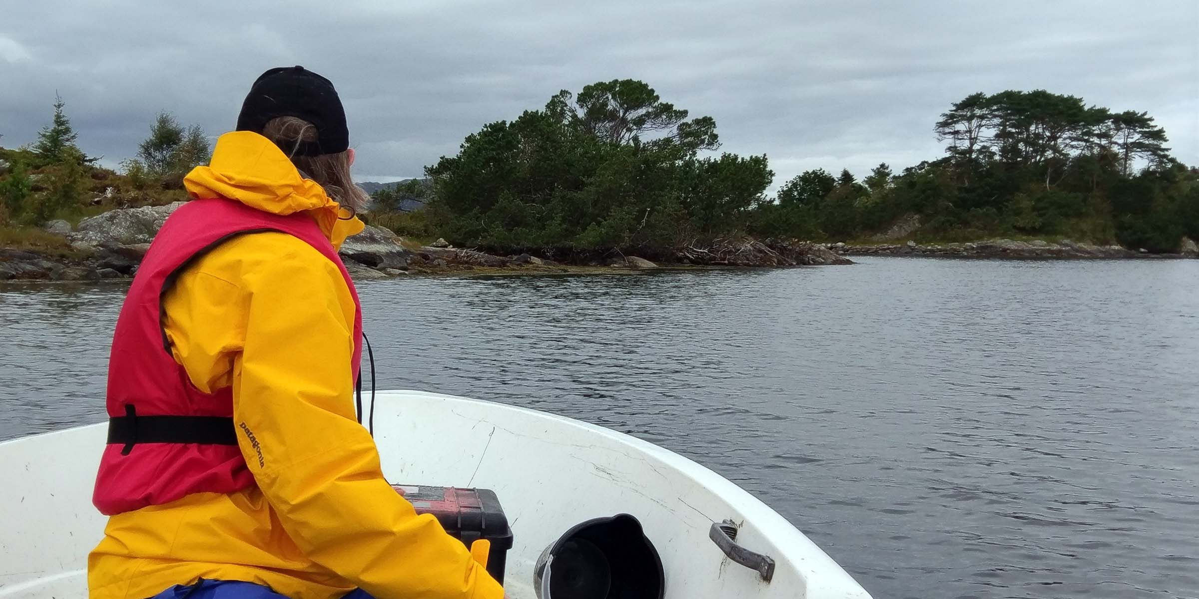 A woman wearing a yellow jacket and red life vest looking forward off the bow of a boat toward rocky shoreline.