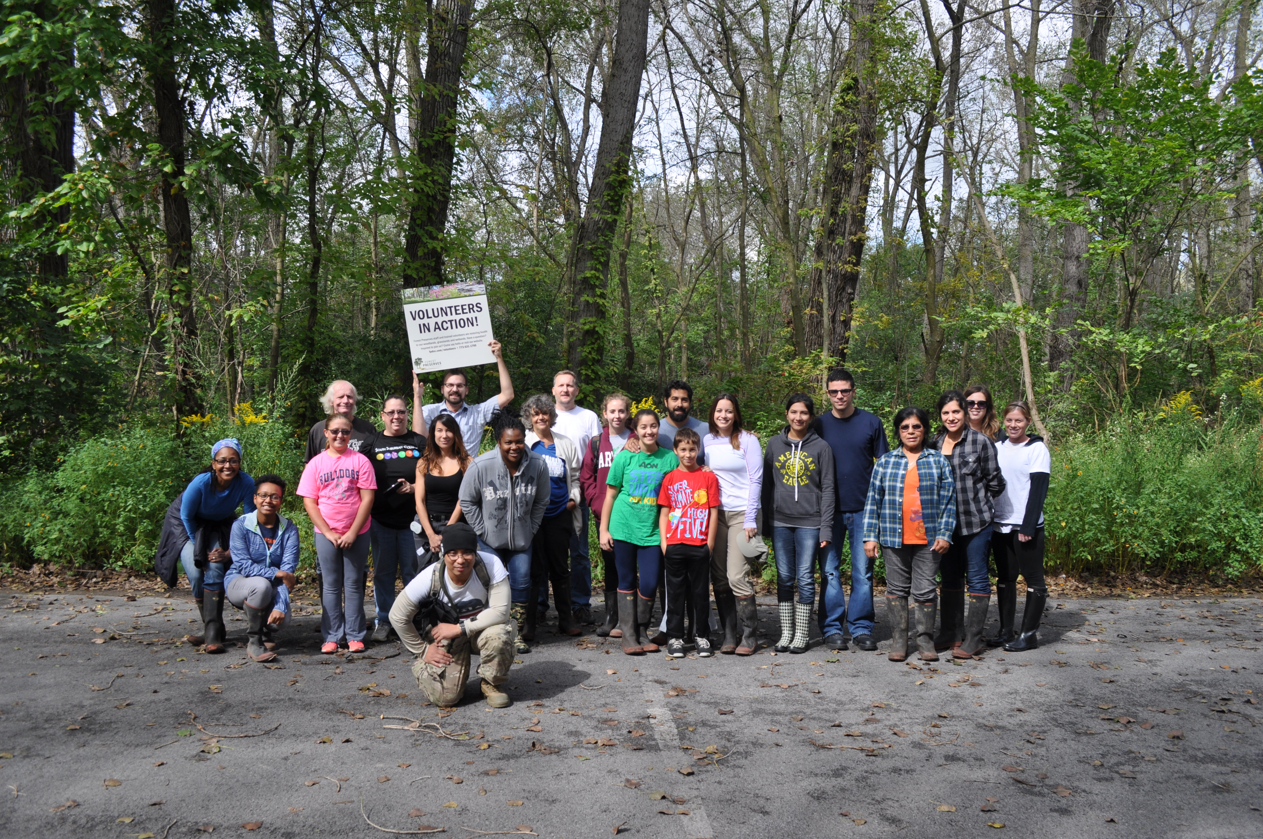 Habitat restoration volunteers of all ages gather for a photo in a wooded area. One person holds up a Volunteers in Action sign.