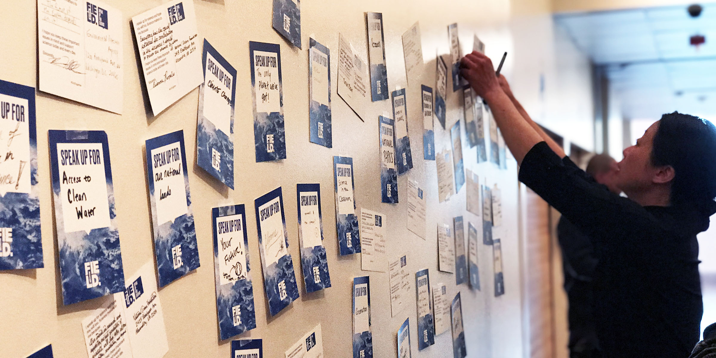 A woman tapes a blue and white postcard to a wall covered in similar postcards.