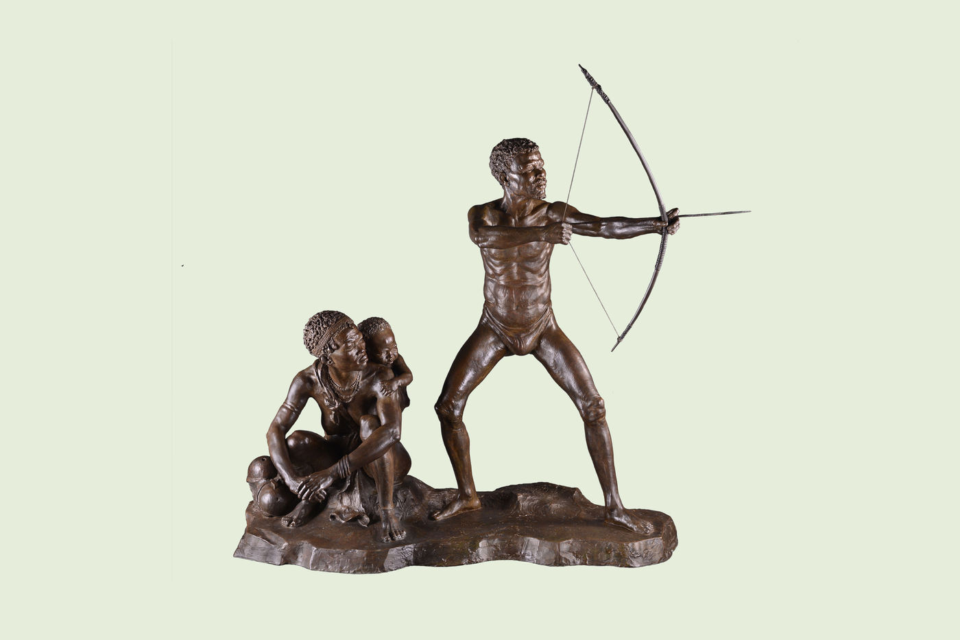A bronze sculpture depicting an adult man standing with a bow and arrow drawn. Next to him, an adult woman sits with a child on her back.