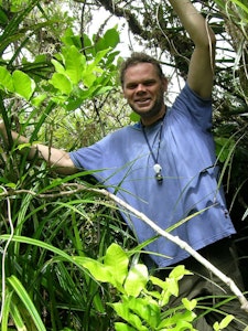 A man in a blue t-shirt smiles while standing among trees and leafy green plants