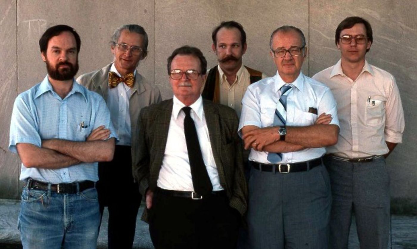 Six men standing side-by-side along a plain concrete wall.