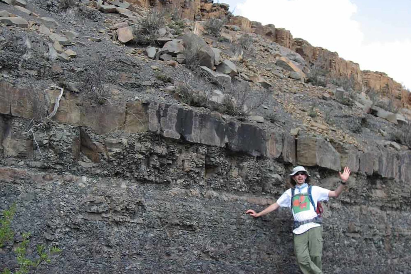 A man wearing a hat, field clothing, and a backpack standing next to a tall hill of exposed rock