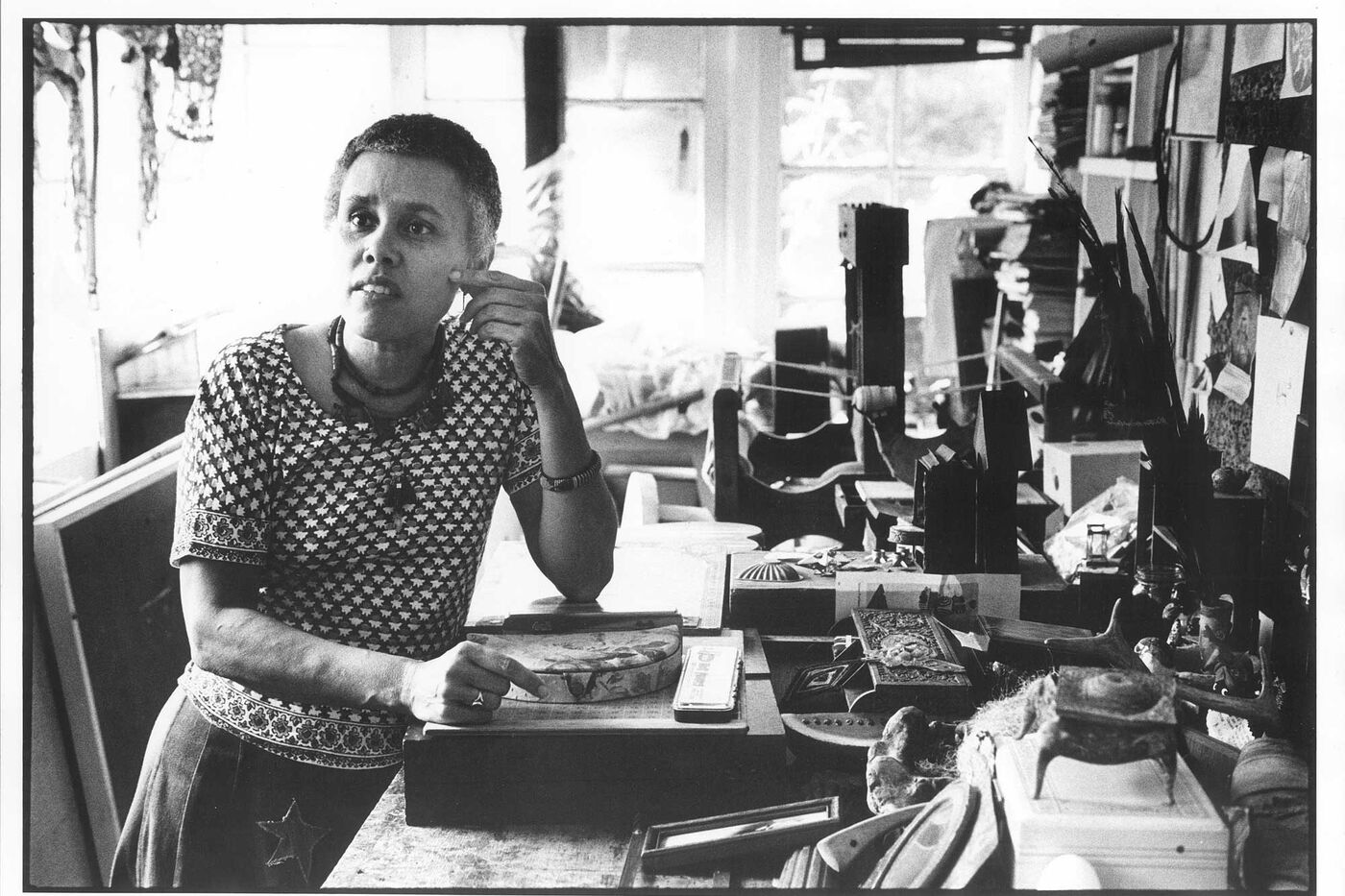 A woman standing while leaning her elbow on a desk. The desk is covered with various wood and metal objects that she uses to make three-dimensional art.