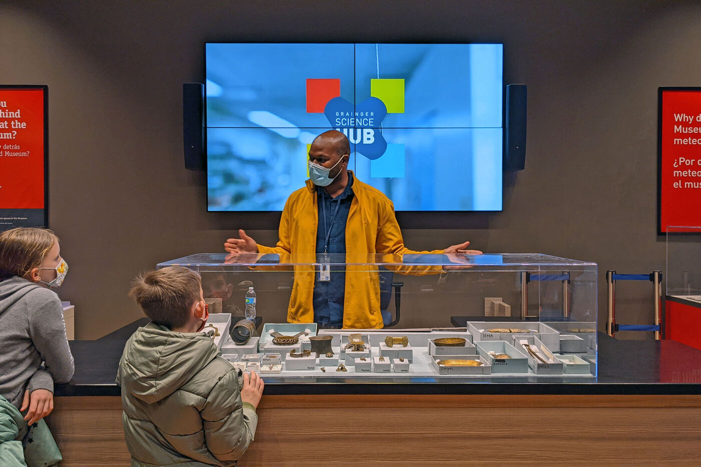 A man stands behind a display case with his arms out-stretched while three museum visitors look on. 