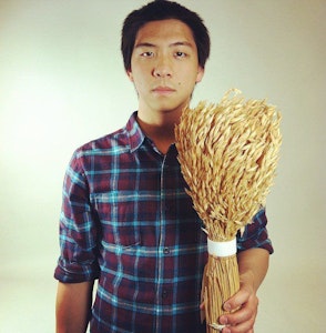 A man in a plaid shirt stands holding a sheaf of wheat