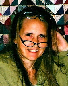 a woman with long hair wears two pairs of eye glasses and smiles in front of a quilted background