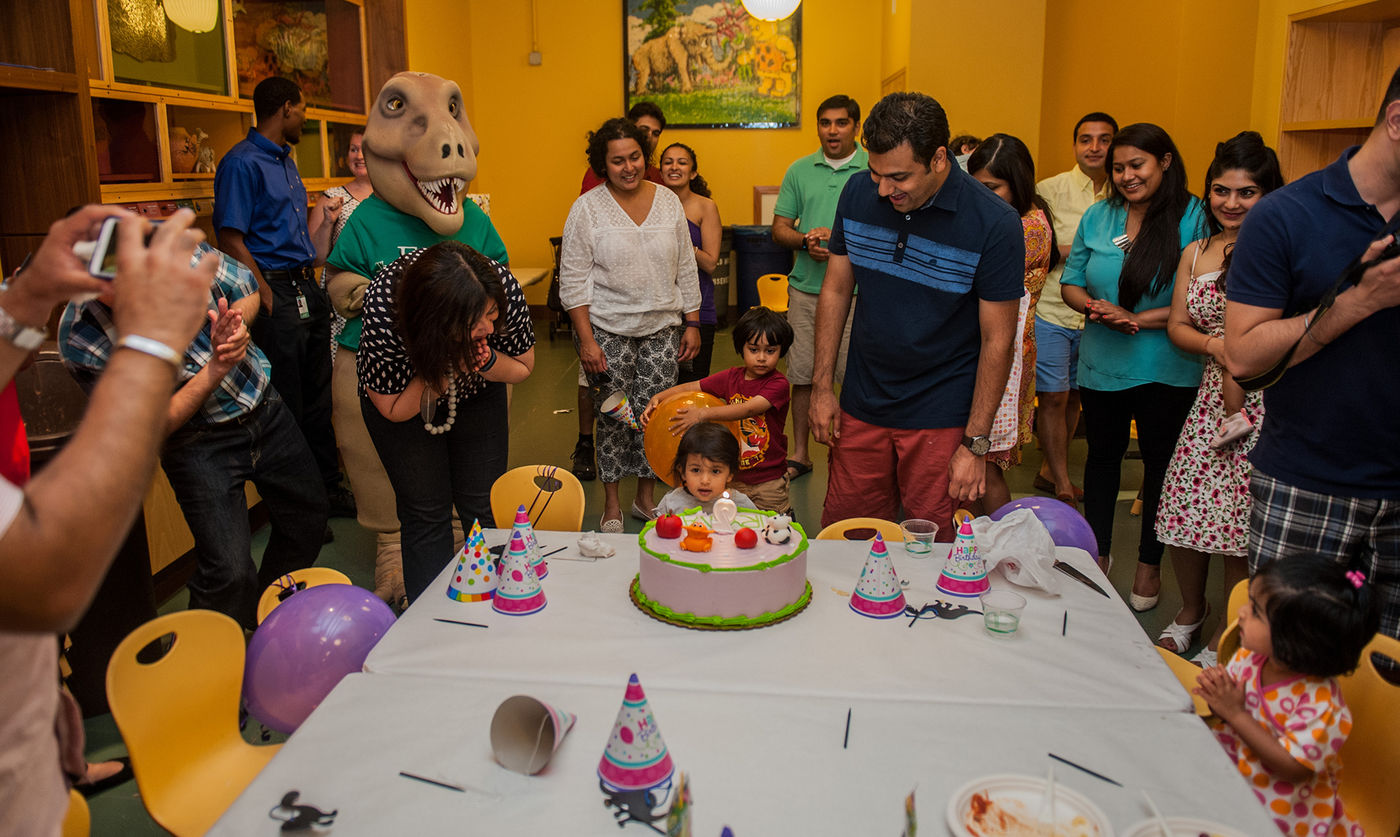 A family celebrates a birthday party in the Art Studio of the Crown Family PlayLab. The group is smiling at a toddler who sits at the end of a table in front of a birthday cake. Cups, napkins, and birthday party hats are scattered on top of the table. A T. rex mascot stands with the crowd. 