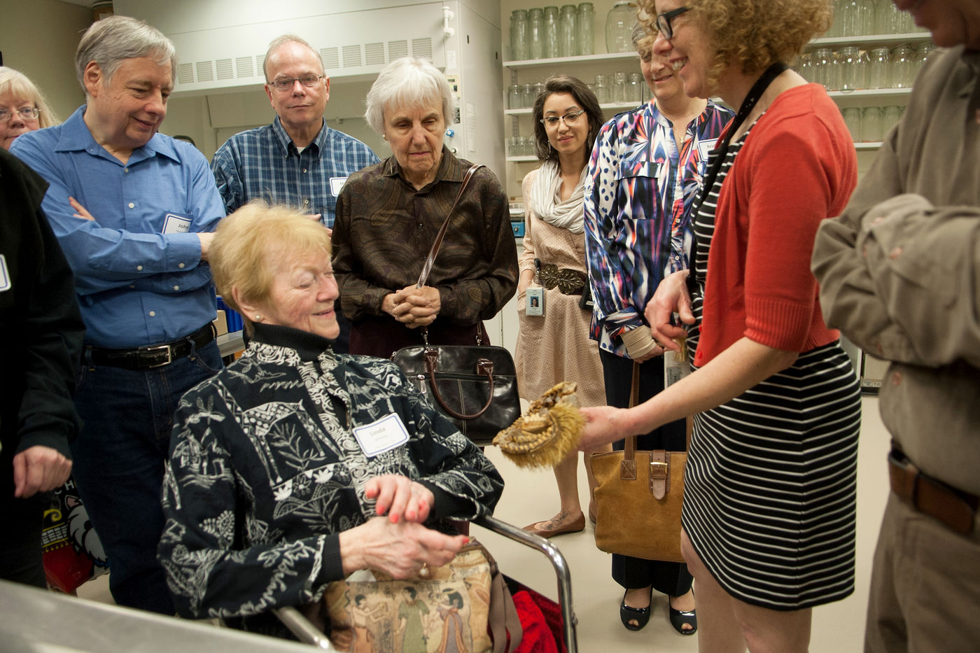 An older woman in a wheelchair smiles as she looks at a specimen held by a staff member. Other adults in the group stand around the pair, looking intently at the specimen. Clear jars are on shelves in the background.