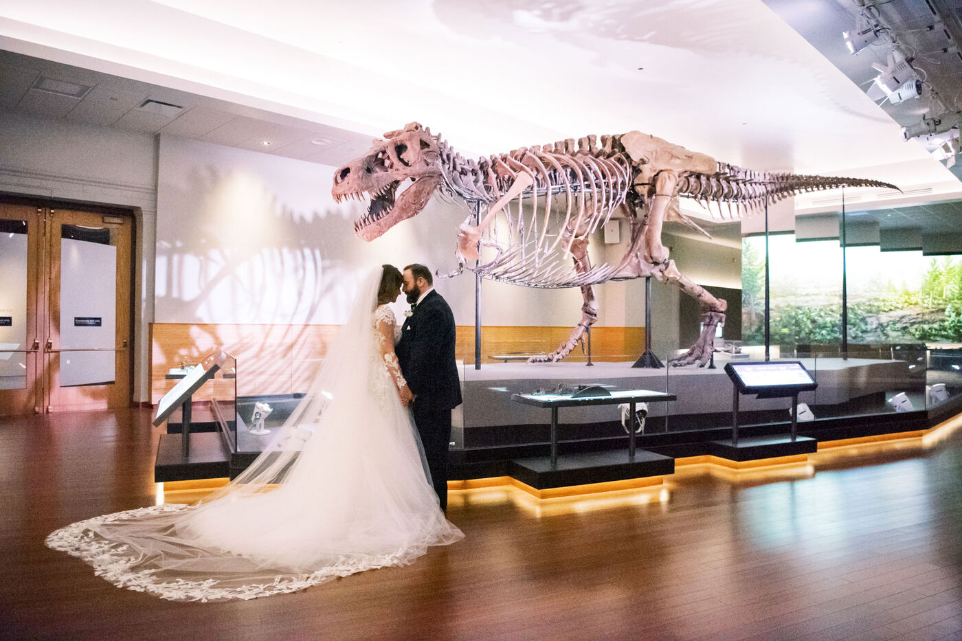 A bride and groom stand in a museum gallery with a T. rex fossil skeleton