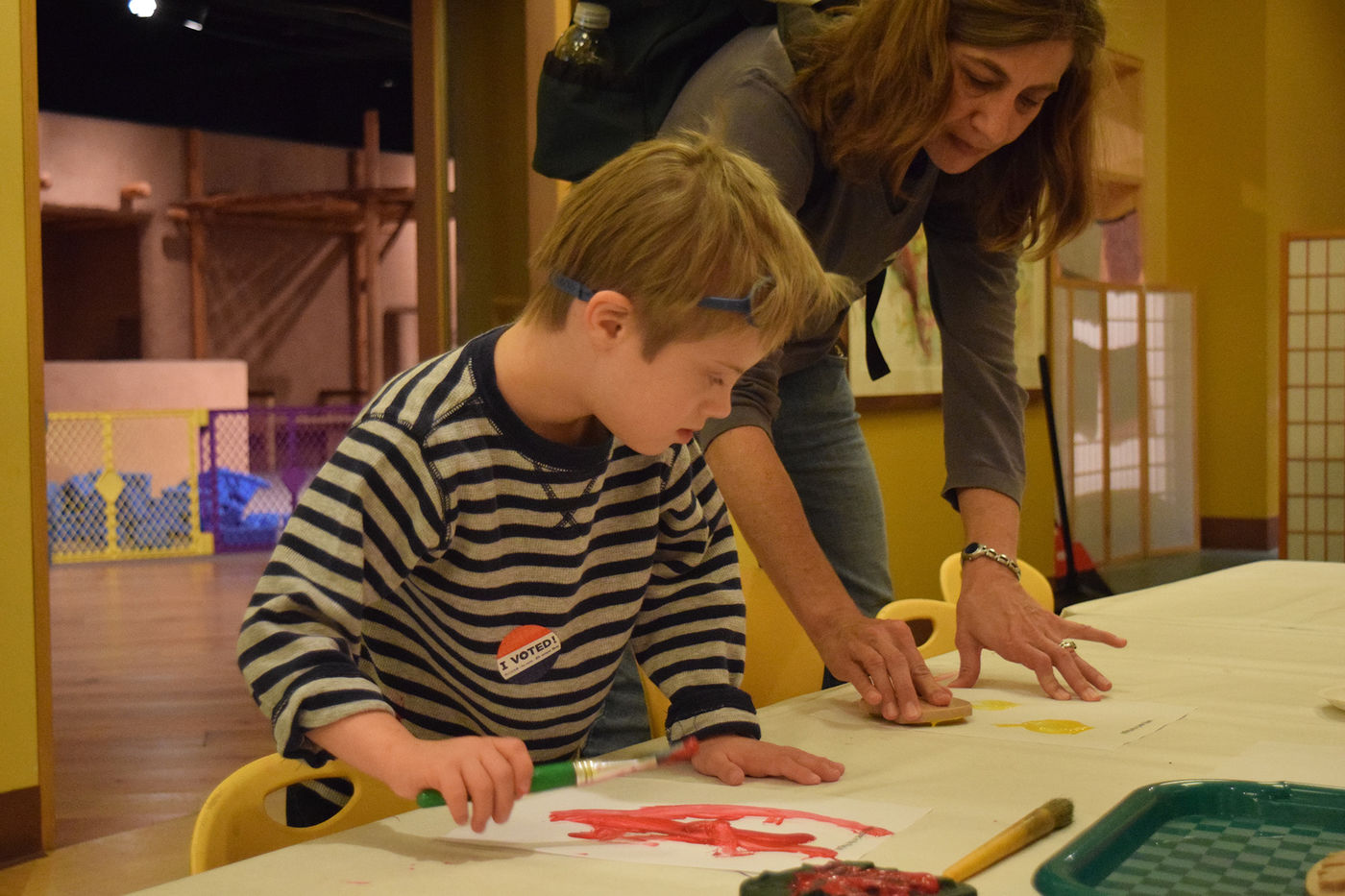 A young visitor paints with his caretaker at a table.