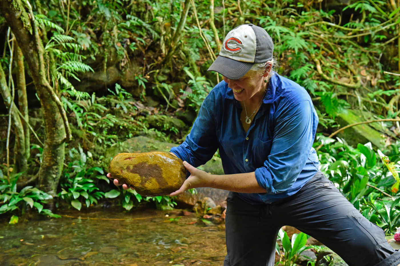 A woman smiles as she stands next to a stream and looks at a large rock. She is in the jungle, surrounded by trees, vines, and bright green leaves.