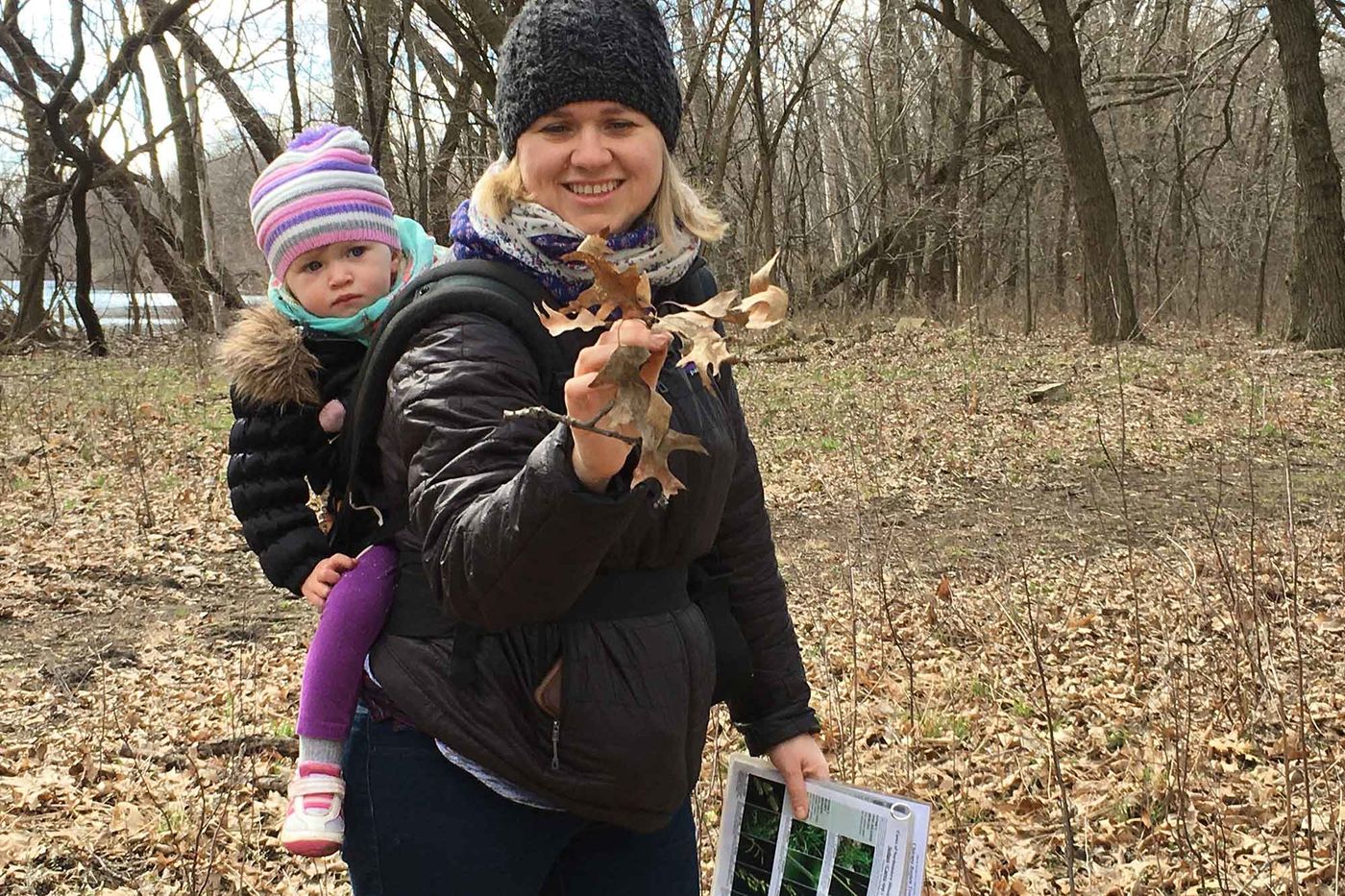 A mother and her young child explore the woods with a Field Museum field guide. The child, attached to her mother's back, looks on as her mother holds a bundle of dried leaves. 