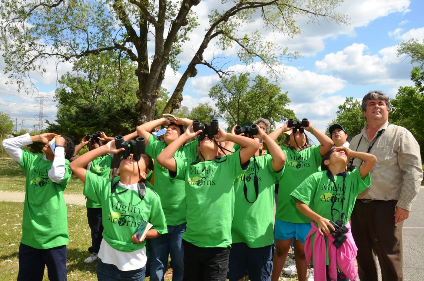 A group of students in matching Mighty Acorns t-shirts use binoculars, looking upwards.
