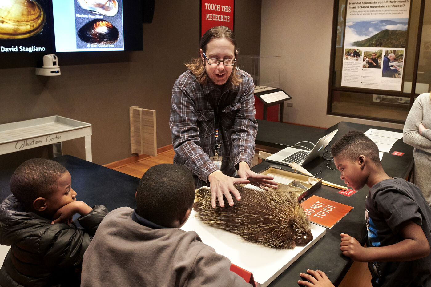 Eli Suzukovich shares a taxidermy specimen with a group of young visitors. The specimen is set on top a table with the visitors gathered around the specimen on the other side of the table. 