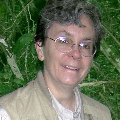 A women with short gray hair and glasses smiles in front of a leafy forest backdrop