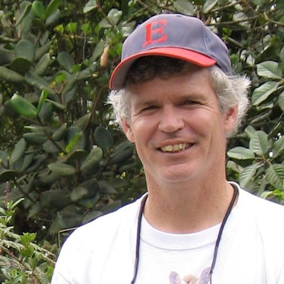 A man with short gray hair and a baseball cap stands in front of green leafy plants