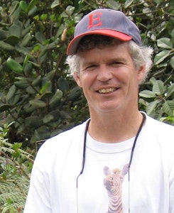A man with short gray hair and a baseball cap stands in front of green leafy plants