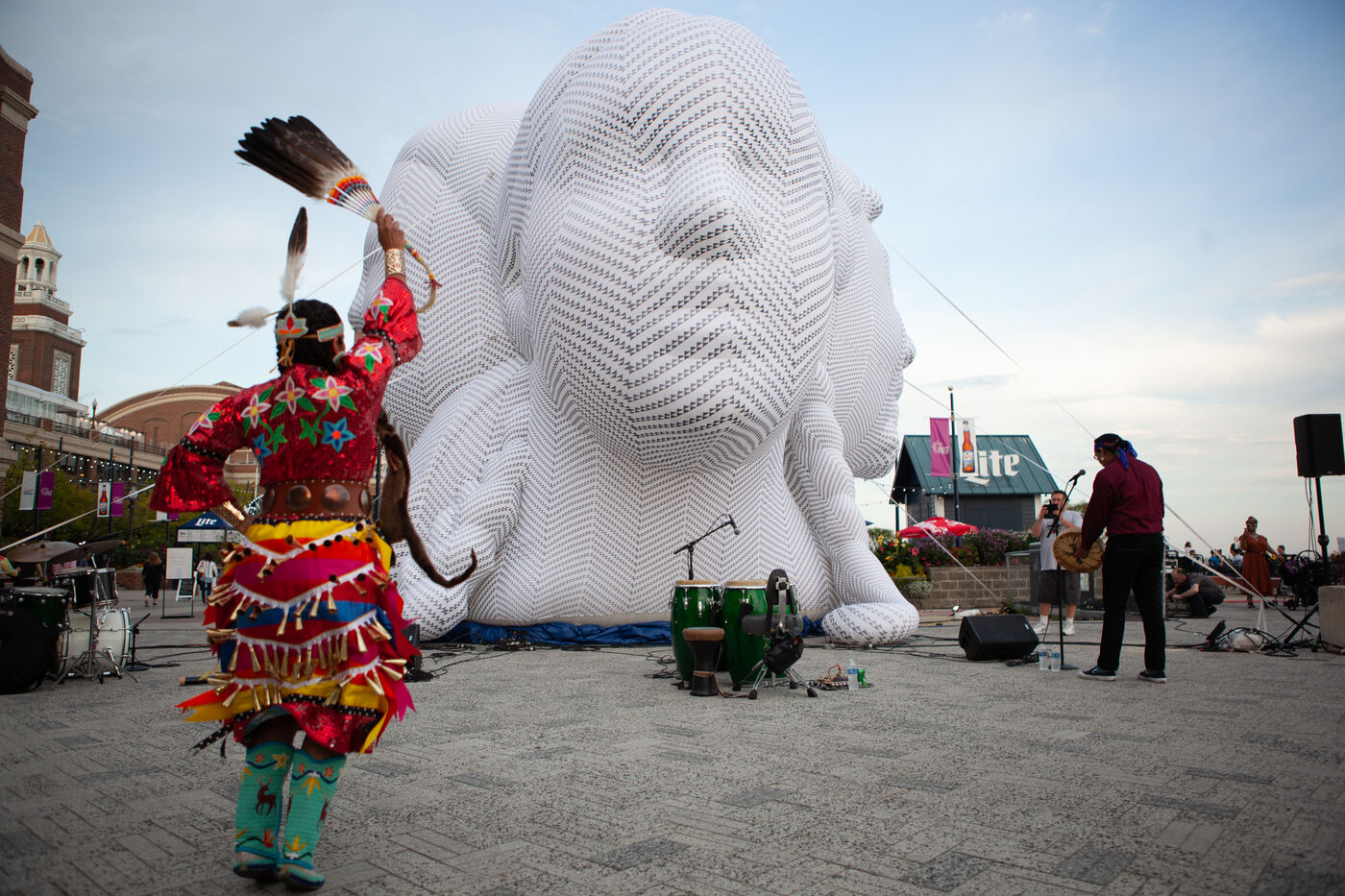 A dancer in traditional dress dances in front of a large outdoor inflatable busts that references DuSable, Kitihawa, Chicago’s first African American Mayor Harold Washington, and a bust of a young boy by artist William Artis. 