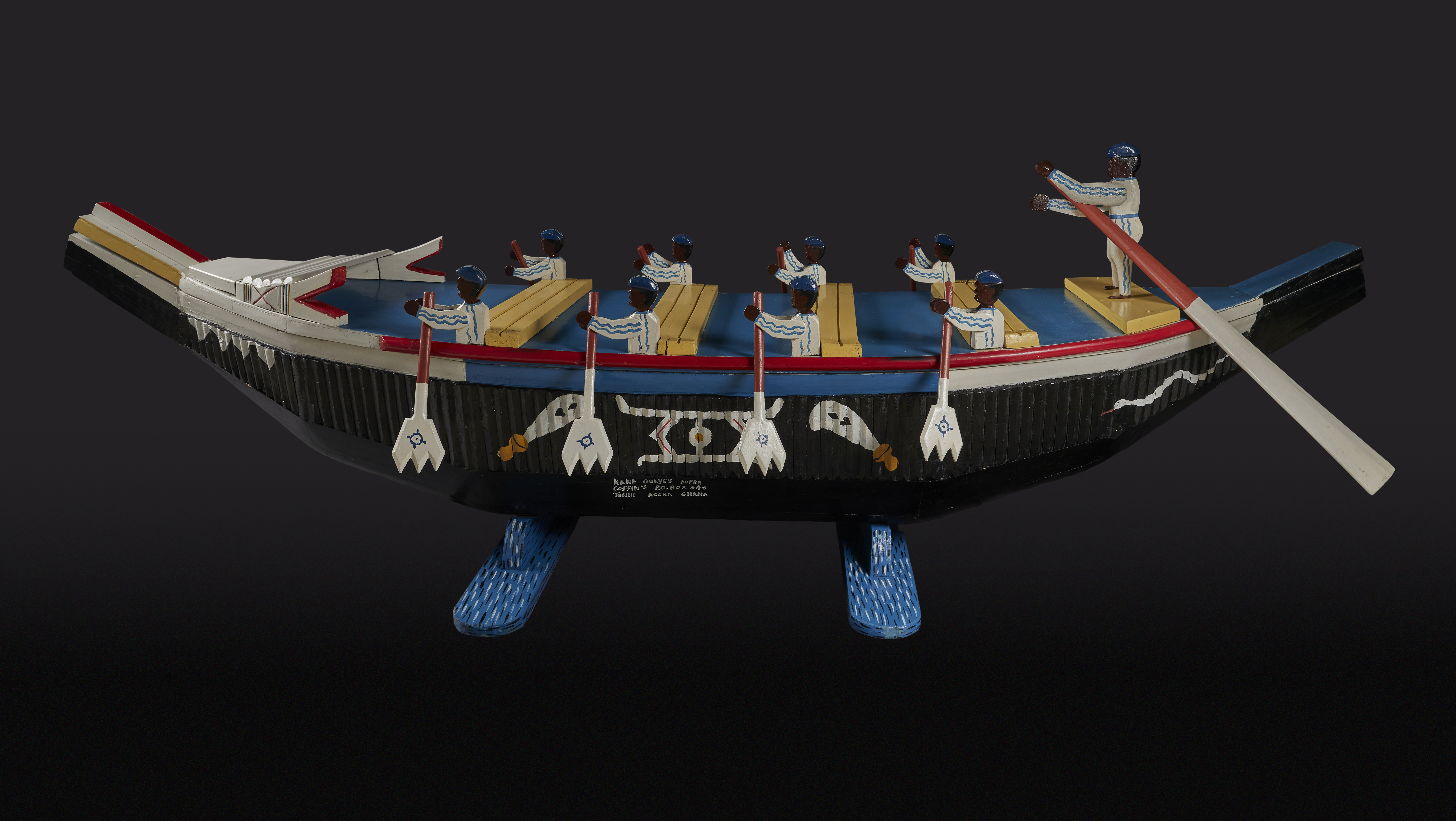A nine-foot long canoe-shaped coffin made of painted wood. The decorations on the coffin depict nine people rowing a black and blue canoe.