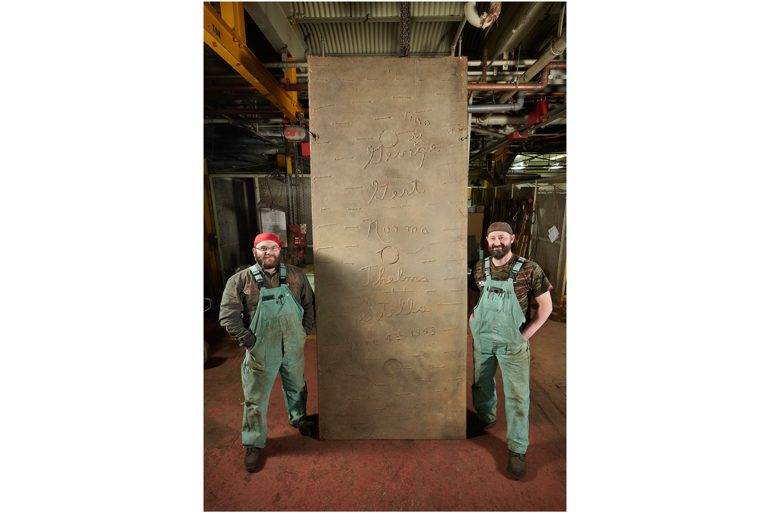 Two steel mill workers in green coveralls pose on either side of a ten-foot-tall steel plate that was created during World War II. The plate features women’s names written in cursive.