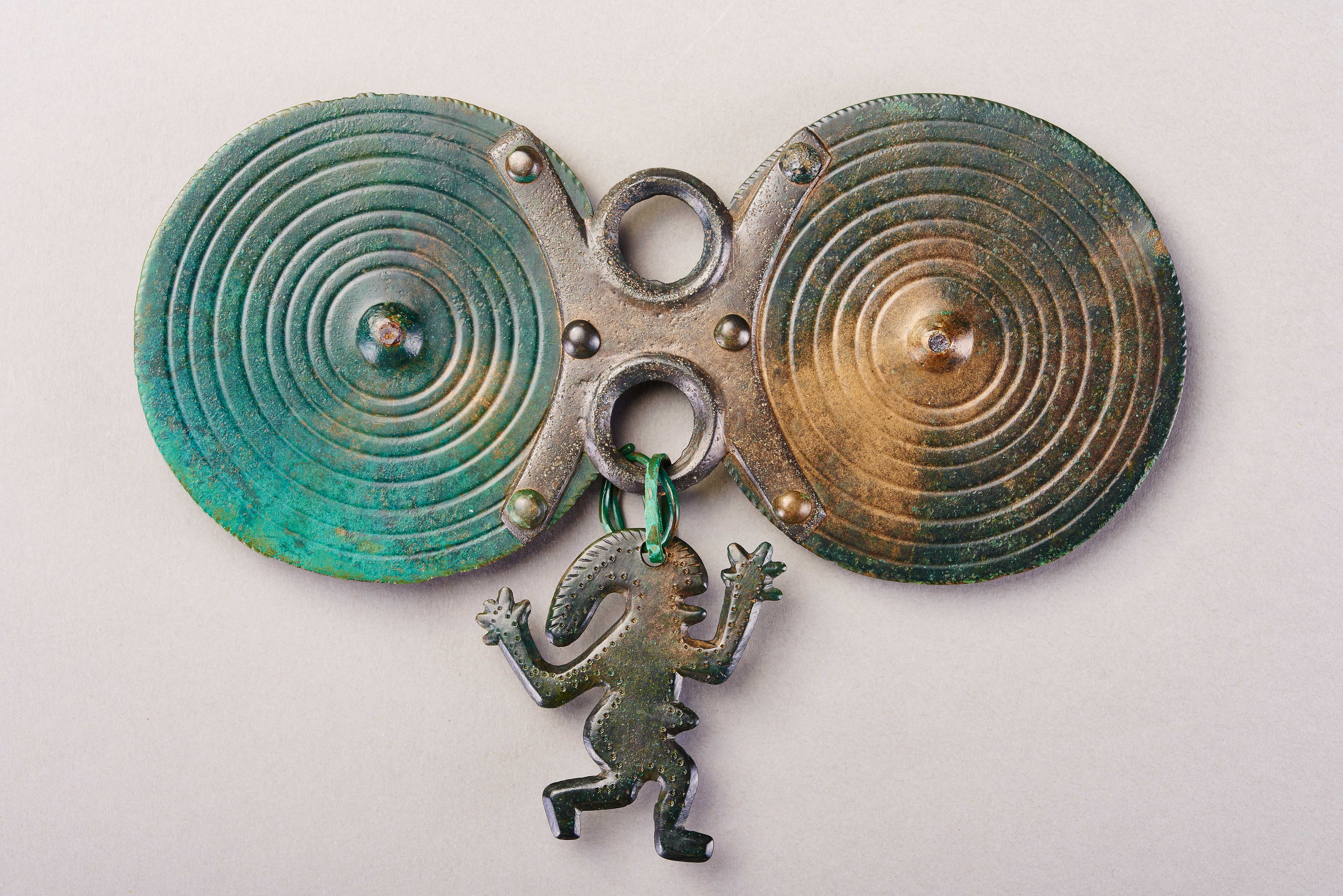 A bronze pin with a green patina. Two circles joined together by two smaller rings, with a bronze figure handing from the middle.