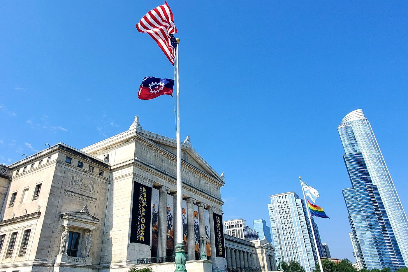 The north facade of the Field Museum, with a clear blue sky and a few high rise buildings in the background. Two flag poles are visible on the museum terraces, one with the United States flag and the Juneteenth flag and the other with the Chicago flag and Pride flag.
